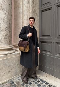 Tumšs mētelis, melns džemperis un plata brūna bikses. Brūna un pelēka soma ar metāla detalēm, nēsāta pāri plecam. Strukturēta akmens fona.