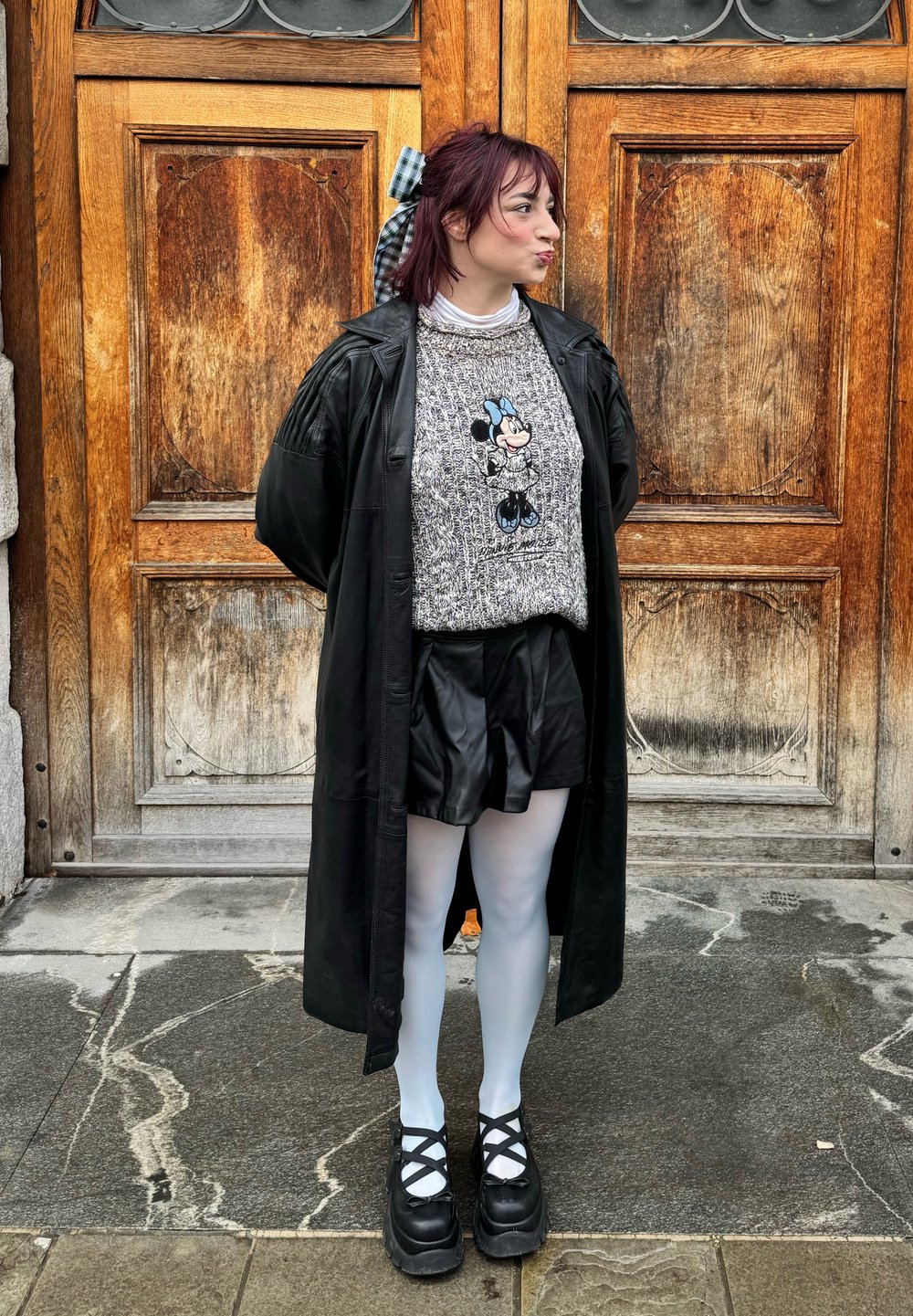 Cappotto lungo nero sopra un maglione grigio con design di Topolino, abbinato a pantaloni corti in finta pelle nera, collant bianchi e scarpe nere chunky.
