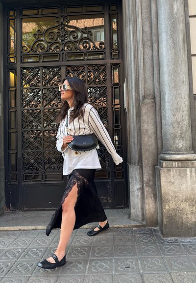 Camisa blanca de botones con rayas negras, falda maxi de encaje negra con abertura, zapatos planos negros y un bolso negro, con un fondo de una puerta de hierro ornamentada.