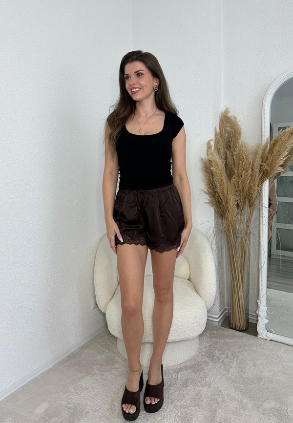 Femme debout dans un haut noir et un short en satin marron avec bordure en dentelle, à côté d'une chaise blanche incurvée et d'une grande herbe de la pampa séchée.