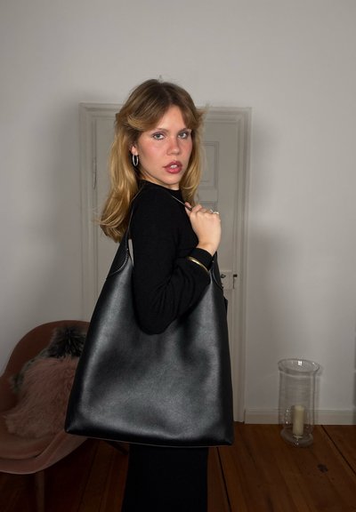 Bolso tote de cuero negro con textura suave, forma grande y asas resistentes. Colocado sobre un fondo neutro que realza el diseño del bolso.