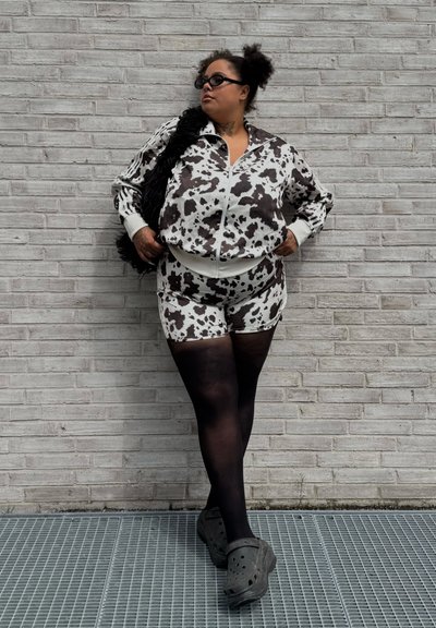 Conjunto de ropa deportiva con un patrón de estampado de vaca en blanco y negro, combinado con mallas transparentes negras y zuecos grises, frente a una pared de ladrillo.