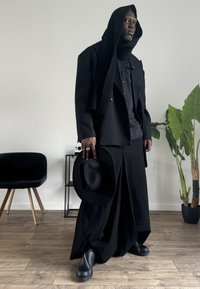 Blazer preto de abotoamento duplo, camisa preta, calças largas pretas, cachecol preto, chapéu preto e calçado preto, com uma planta em vaso ao fundo.