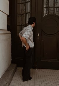 Giovane uomo con camicia a quadri e pantaloni neri si trova di fronte a una grande porta in legno scuro con pannelli di vetro, all'interno.