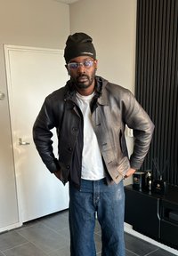 Veste en cuir marron avec fermeture à boutons, poignets côtelés et capuche. Associée à un t-shirt blanc et un jean bleu. Lunettes transparentes et bonnet noir.