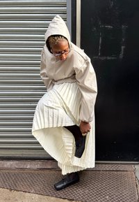 Lichtgrijs gewatteerd jack met capuchon en geplooide mouwen, gecombineerd met een lange witte geplooide rok en zwarte veterlaarzen op een gestructureerde ondergrond.