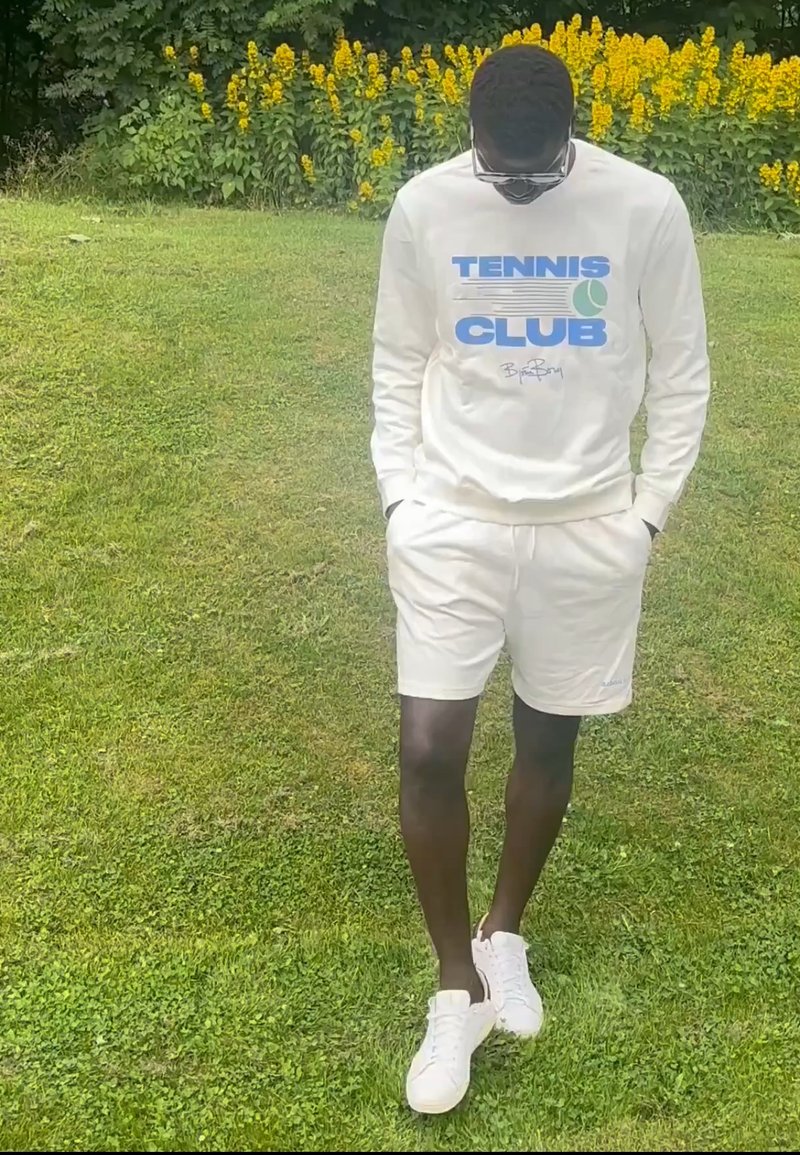 Homme portant un sweat-shirt blanc du club de tennis, un short blanc et des chaussures blanches, debout sur l'herbe avec des fleurs jaunes en arrière-plan, regardant vers le bas.
