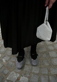 Μαύρη πλισέ φούστα, μαύρες καλσόν, στολισμένα μαύρα παπούτσια με ασημένιους κοσμήτες και μια μικρή ασημένια τσάντα από χάντρες. Πλακόστρωτη επιφάνεια από κάτω.