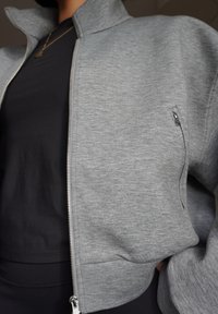 Grey zip-up jacket made of soft material, featuring a high collar and a side pocket with a silver zipper. Worn over a black top.