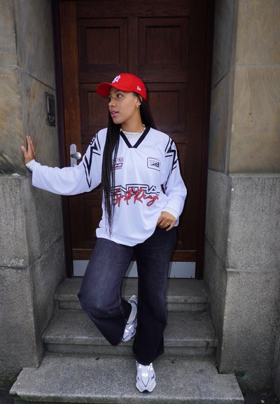 Jersey de hockey de manga larga blanca con detalles en negro, jeans oscuros de gran tamaño, gorra roja y zapatillas plateadas, de pie en unos escalones de piedra frente a una puerta de madera.