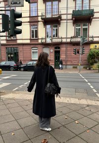 Fekete kabátot és szürke nadrágot viselő személy vár a gyalogátkelőhelyen a piros lámpánál, a háttérben történelmi épület és parkoló autók láthatók.