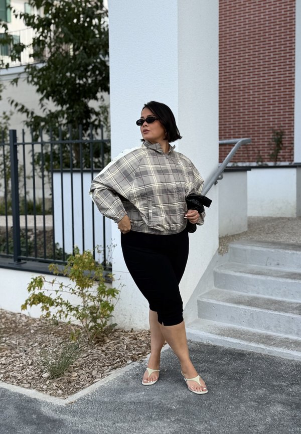 Femme portant des lunettes de soleil, une veste à carreaux, un pantalon capri noir et des sandales, debout près d'un escalier et d'un bâtiment blanc avec des plantes et une rambarde à proximité.