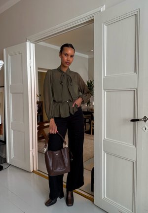 Blusa verde oliva con lazo, pantalones negros y bolso de mano marrón oscuro. Botas de tacón negras con superficie texturizada. Fondo interior claro.