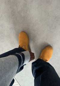 Person som har på seg brune arbeidsstøvler, mørke jeans og en grå genser med marineblå striper, står på et lysegrått gulv.