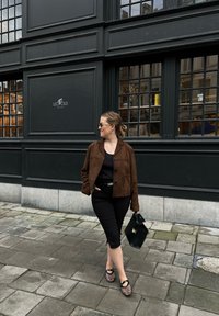 Veste en daim marron, haut noir, pantalon noir ajusté, ballerines à motifs et sac à main noir, se tenant sur des pavés près d'une façade de bâtiment sombre.