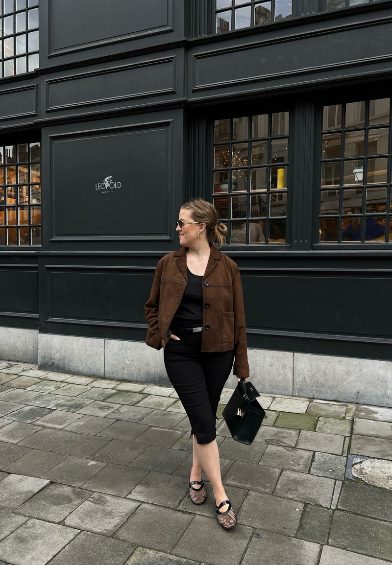 Καφέ σουέντ μπουφάν, μαύρη μπλούζα, μαύρο εφαρμοστό παντελόνι, σχέδιο παπούτσια και μαύρη τσάντα, στέκεται σε κυβόλιθους κοντά σε μια σκοτεινή πρόσοψη κτηρίου.