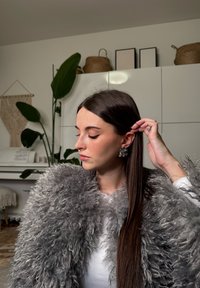 Femme aux longs cheveux bruns, portant un manteau gris duveteux et de grandes boucles d'oreilles argentées en forme de feuille, ajustant une boucle dans un cadre intérieur moderne.