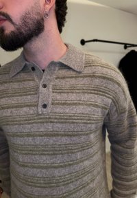 Homme avec une barbe courte et des boucles d'oreilles doubles en forme d'anneaux, portant un pull en maille rayé gris et olive avec un col et trois boutons.