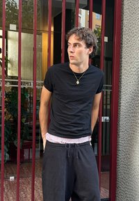 Mladý muž v čiernom tričku a nohaviciach stojí s rukami v vreckách, pozerajúc doľava, pred červenými kovovými tyčami a sklenenými dverami.