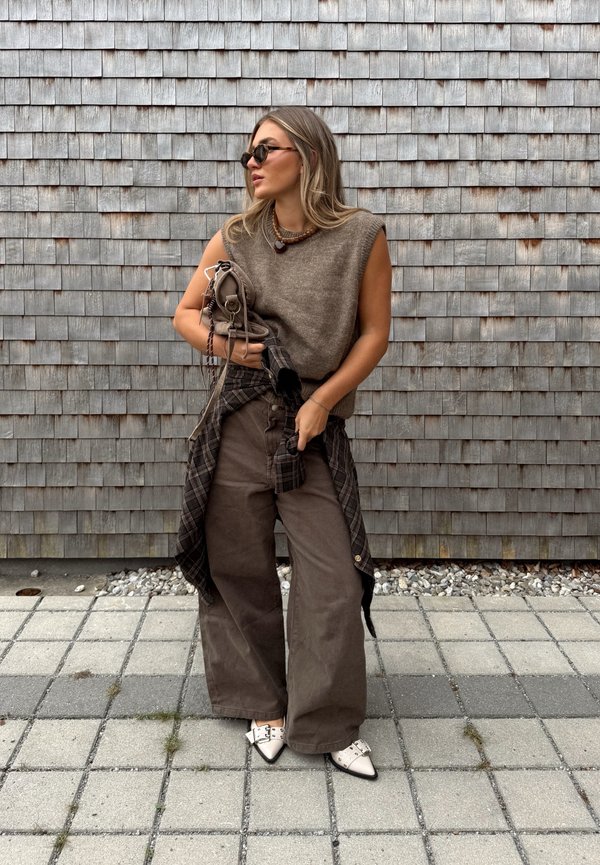 Pull en tricot sans manches beige, pantalon large marron, chemise à carreaux noire nouée à la taille, mocassins blancs à bout pointu avec quincaillerie argentée, arrière-plan en bois gris.