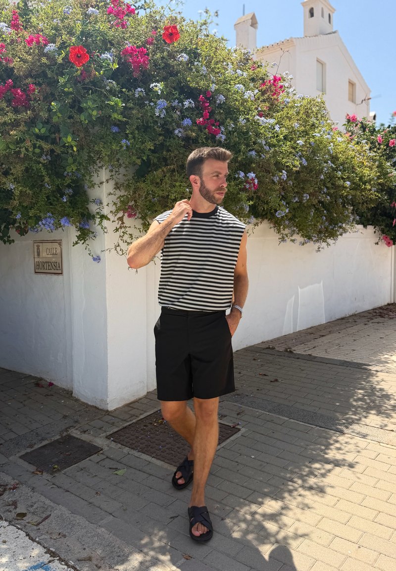 Uomo con pantaloncini neri e maglietta senza maniche a righe che cammina per una strada soleggiata accanto a un muro bianco con cespugli in fiore e un cartello stradale con scritto Calle Hortensia.