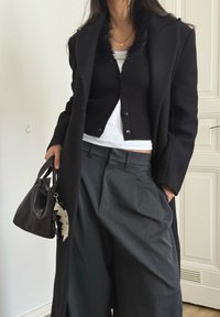 Black wool coat with a faux fur collar, worn over a black ribbed cardigan and a white striped cropped top. Grey high-waisted trousers and a black handbag.