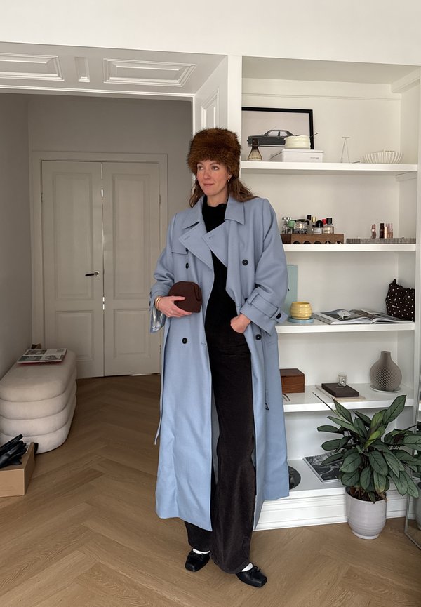 Vrouw in een lichtblauwe trenchcoat en een bruine bontmuts, met een bruine clutch in haar hand, staand in een kamer met witte planken en een houten vloer.