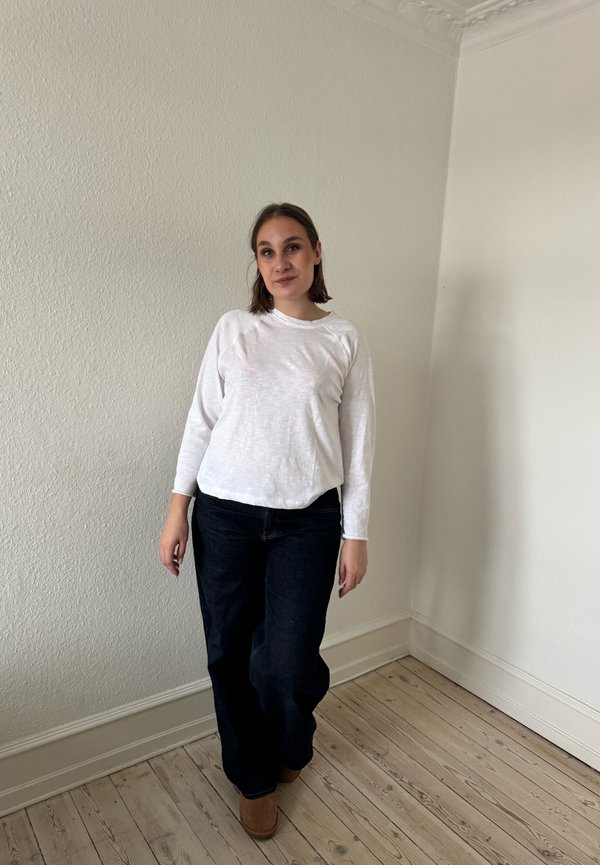 Žena s hnedými vlasmi dĺžky po ramená, oblečená v bielej tričku s dlhým rukávom, tmavých džínsoch a hnedých topánkach stojí v prázdnej izbe v kúte s drevenou podlahou.