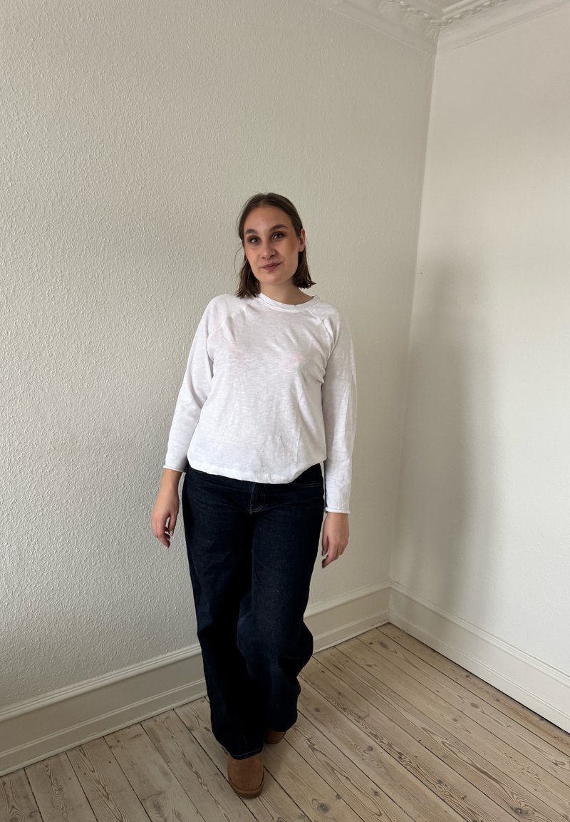 Femme aux cheveux bruns mi-longs, portant un chemisier blanc à manches longues, un jean foncé et des chaussures marron, se tient dans un coin vide d'une pièce avec un sol en bois.