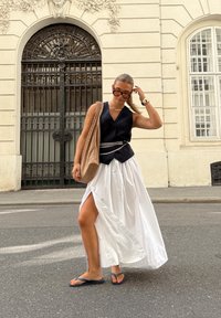 Schwarzes ärmelloses Top, weißer Rock mit seitlichem Schlitz, geflochtene beigen Tragetasche und Flip-Flops. Der Hintergrund zeigt eine verzierte Tür und Wand.