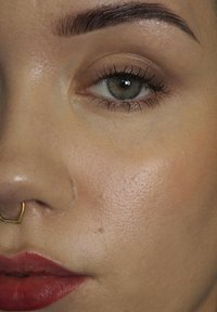 Close-up van een gezicht met lichte huid, een gouden septum piercing, volle lippen met rode lipstick en een gehighlighted huid die een gladde textuur creëert.