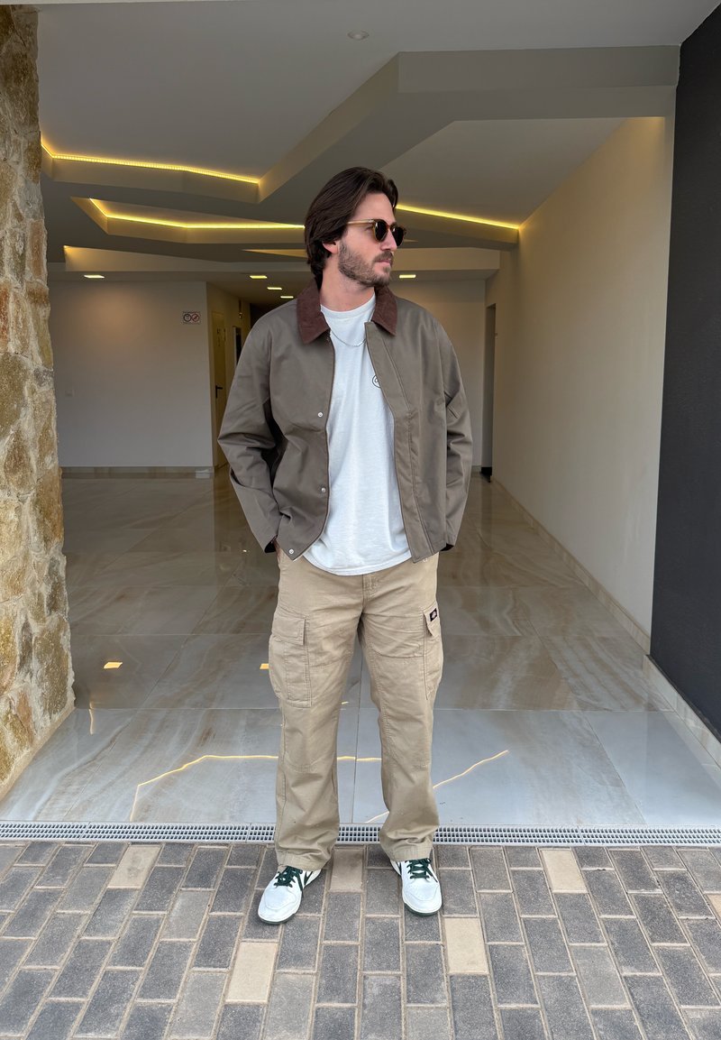 Veste marron et grise avec col, t-shirt gris clair, pantalon cargo beige et baskets blanches avec accents verts, debout sur un sol carrelé.