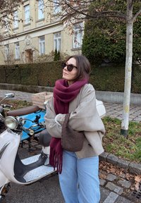 Persoană purtând un pulover gri supradimensionat, eșarfă burgundy și blugi albaștri, stând lângă un scuter alb și o motocicletă albastră.