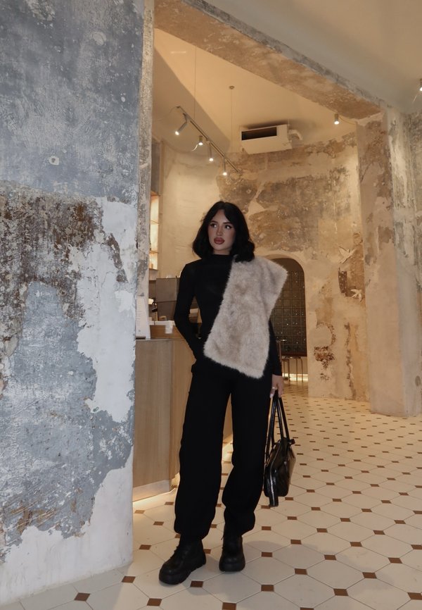 Femme en tenue noire avec étole en fourrure beige, debout dans un intérieur rustique aux murs usés et sol carrelé hexagonal, tenant un sac à main noir.