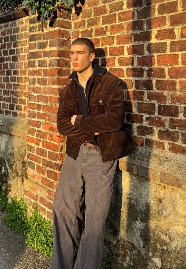 Brązowa kurtka z korduroy z kołnierzem, granatowa koszula na zamek pod spodem i jasnoszare szerokie spodnie; stoi oparta o ceglaną ścianę.