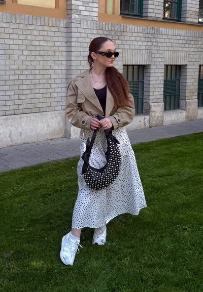 Mujer con el pelo largo que lleva gafas de sol, chaqueta beige, top negro, falda blanca con lunares, zapatillas blancas, sostiene un bolso negro con tachuelas, de pie sobre la hierba.