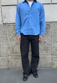 Camicia blu a maniche lunghe con bottoni, abbinata a jeans nero a gamba dritta e scarpe nere, su uno sfondo di muro di pietra strutturato.