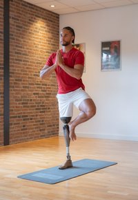 Man die yoga beoefent op één been met een prothese, gekleed in een rode t-shirt en witte shorts, op een grijze yogamat met een bakstenen muur op de achtergrond.