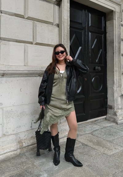 Joven mujer con gafas de sol, chaqueta de cuero negra, vestido verde asimétrico, botas negras, sonriendo y de pie junto a una gran puerta negra sobre un pavimento de piedra.