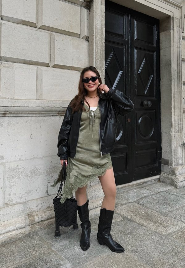 Tânără purtând ochelari de soare, geacă de piele neagră, rochie verde asimetrică, cizme negre, zâmbitoare și stând lângă o ușă mare neagră pe o pavea din piatră.