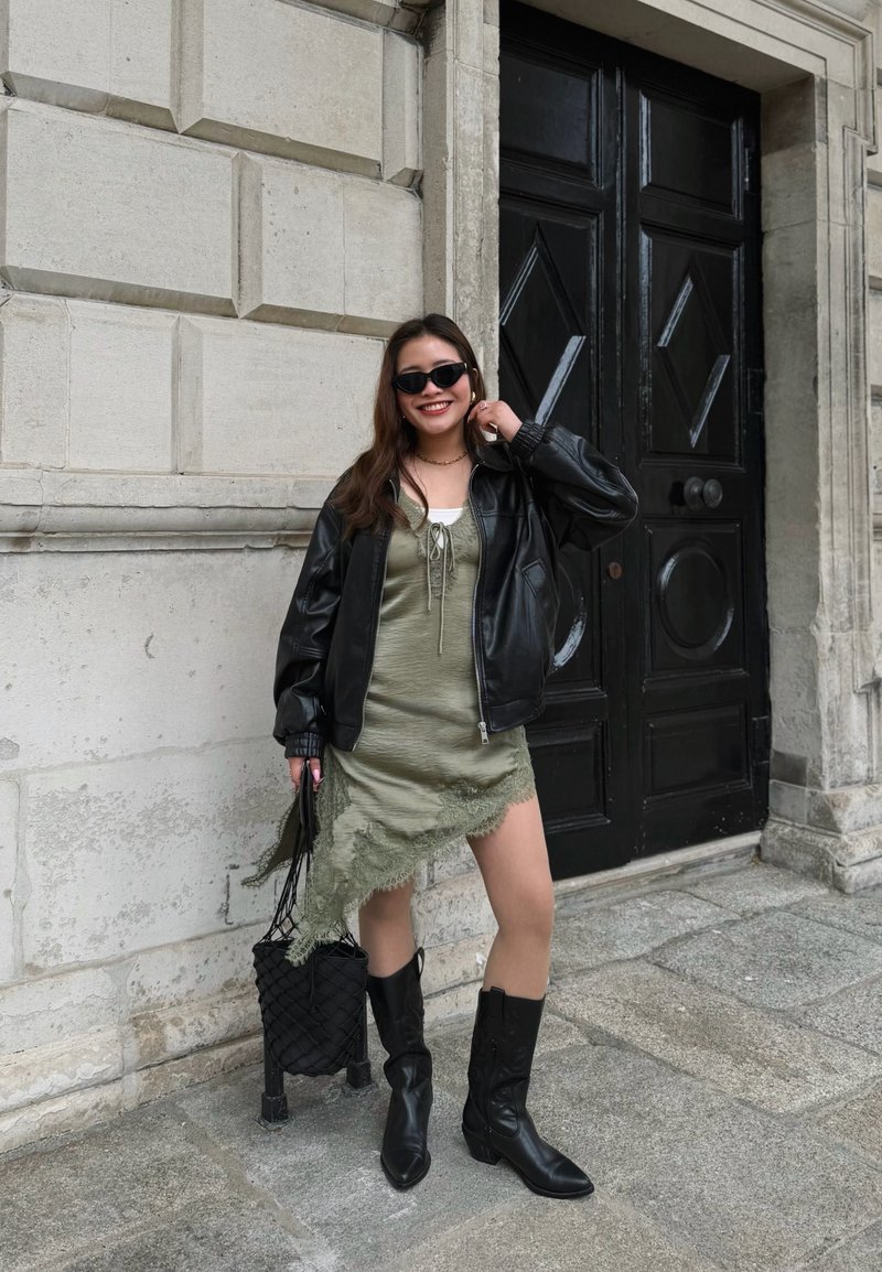 Νεαρή γυναίκα με γυαλιά ηλίου, μαύρο δερμάτινο μπουφάν, πράσινο ασύμμετρο φόρεμα, μαύρες μπότες, χαμογελαστή και στέκεται δίπλα σε μια μεγάλη μαύρη πόρτα σε πέτρινο δάπεδο.