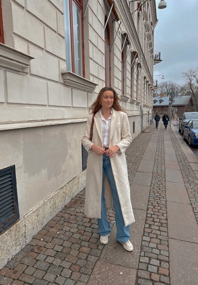 Abrigo largo crema hecho de una tela texturizada, combinado con una camisa blanca y jeans acampanados azules. El sujeto está de pie en una calle empedrada.