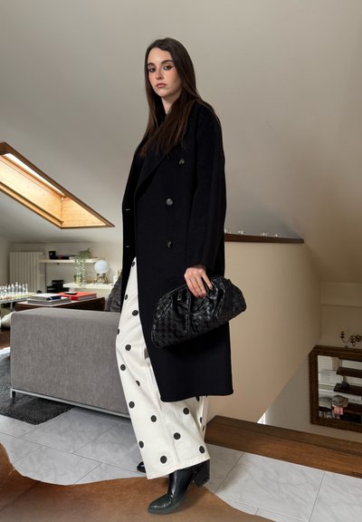 Mujer con abrigo negro, pantalones blancos de pierna ancha con lunares negros, botas negras, sosteniendo un bolso de mano negro tejido en una sala de estar moderna.