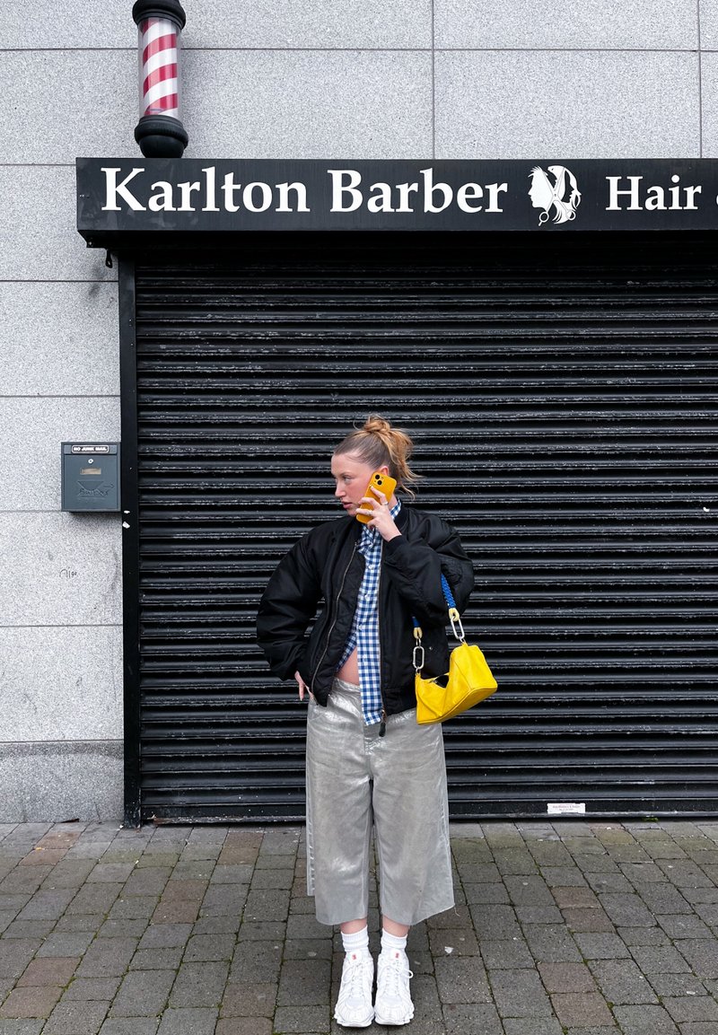 Femme en pantalon argenté et veste noire parle au téléphone devant le salon de coiffure Karlton fermé avec un poteau de barbier rayé au-dessus.