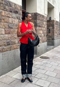Raudona berankovė palaidinė su mygtukų detale, tamsiai mėlyni džinsai su prisiūtu kraštu, ir juoda petinė rankinė. Lygūs audiniai, kasdienis dizainas.