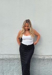 Sort midiskjørt med polkadotter, tettsittende i midjen, kombinert med en hvit tanktopp. Langt, bølget blondt hår. Står foran en lys grå vegg.