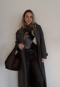 Long manteau en laine gris, pull à motifs noir et blanc, pantalon noir taille haute, sac à main tressé marron, accessoires minimalistes, texture subtile.