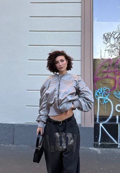 Mujer de cabello rizado con chaqueta bomber plateada y pantalones anchos negros con tachuelas que forman la palabra "GLORY", sosteniendo un bolso negro, de pie junto a una pared cubierta de grafitis.