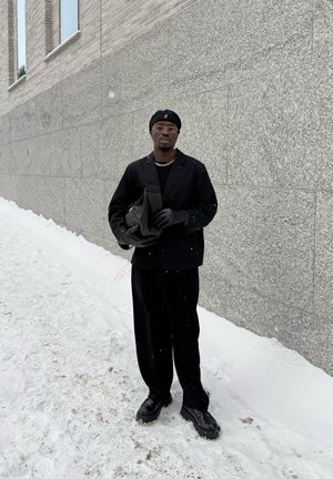 Hombre con boina negra, gafas, guantes y traje sosteniendo una bolsa negra, de pie sobre suelo nevado junto a una pared de piedra gris.