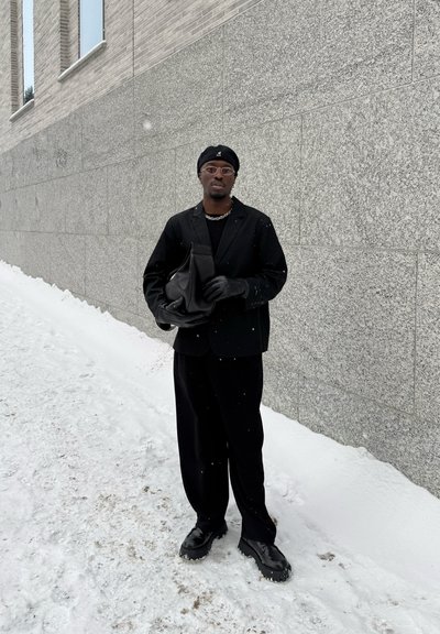 Hombre con boina negra, gafas, guantes y traje sosteniendo una bolsa negra, de pie sobre suelo nevado junto a una pared de piedra gris.